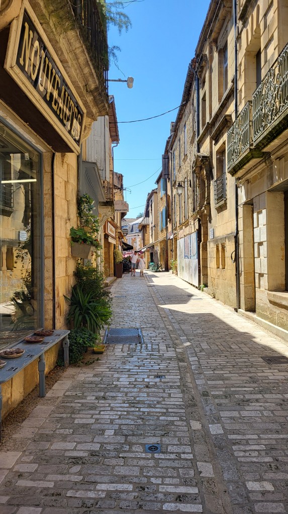 Cobblestone street in Belvès lined with medieval stone houses under soft early evening light.