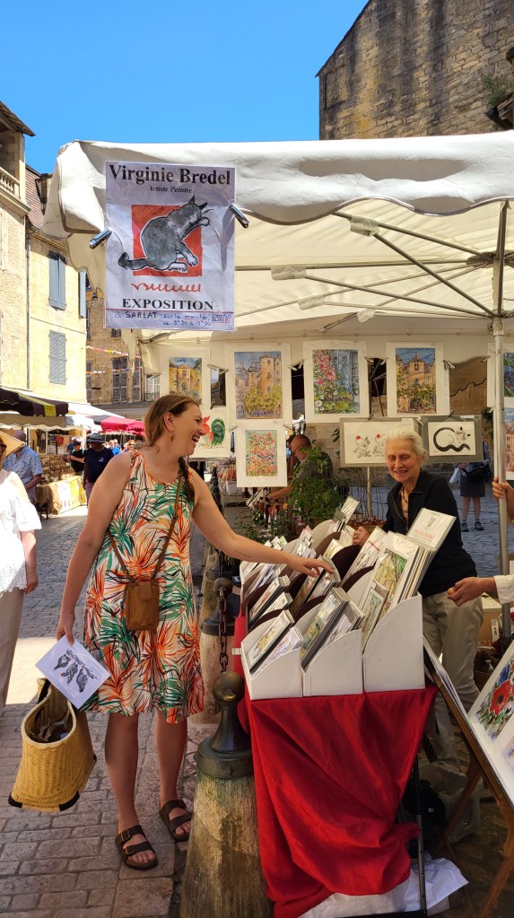 Outdoor art market in Sarlat-la-Canéda featuring Virginie Bredel’s stall with colorful prints, a tropical-dressed visitor, and historic stone buildings under a summer sky