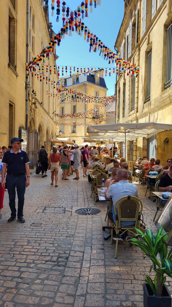 Festive street scene in Sarlat-la-Canéda with colorful pom-pom decorations, historic stone buildings, and locals dining outdoors under a clear summer sky