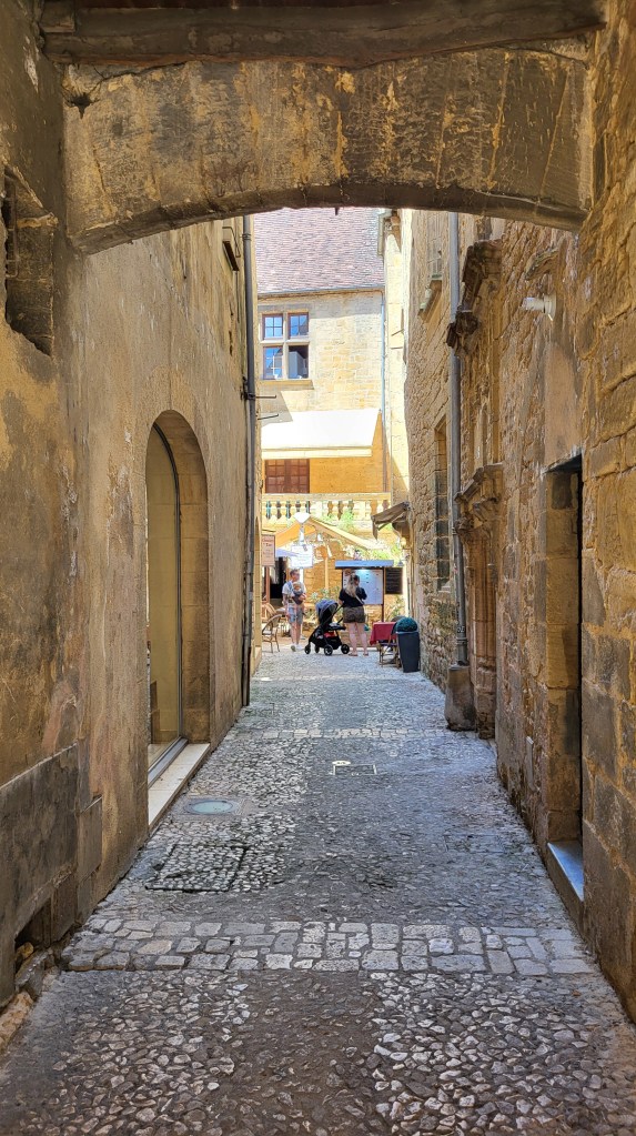 Cobbled alleyway in Sarlat-la-Canéda framed by medieval stone archways, glimpsing market stalls and morning activity in the distance